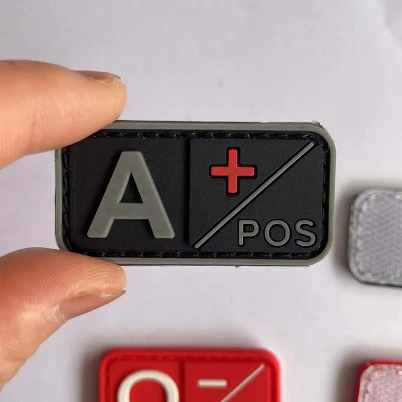 A close-up of a hand holding a rectangular black tactical patch with gray lettering reading 'A' and 'POS' and a red plus sign, on a light gray background with other patches partially visible.
