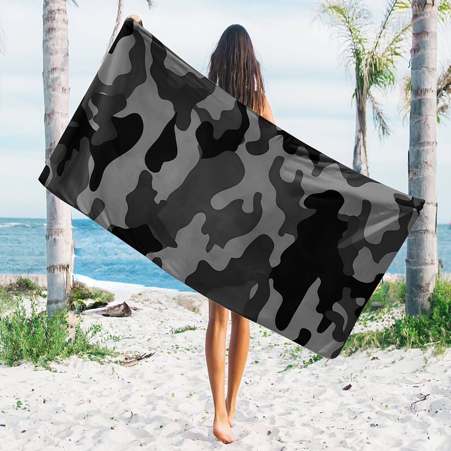 Camouflage beach towel displayed by a person on a sandy beach with palm trees and ocean in the background
