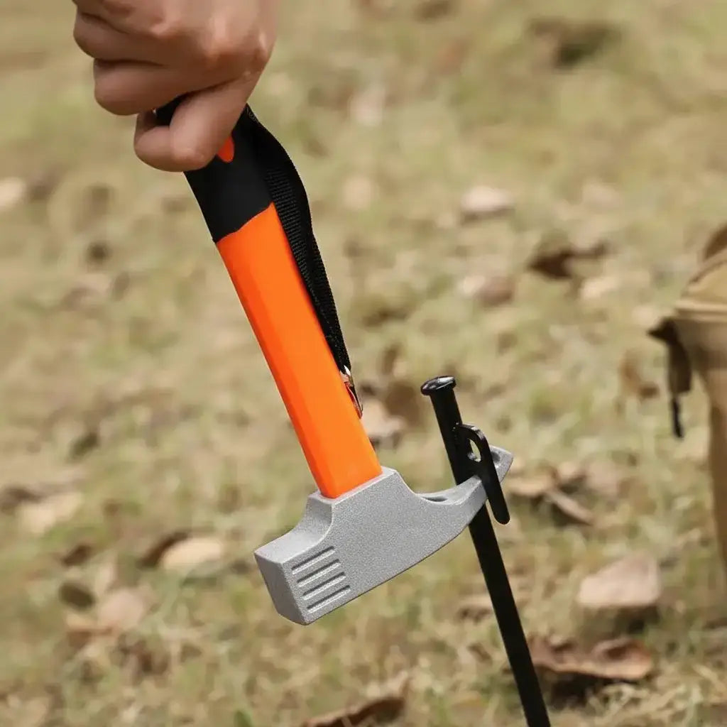 A close-up of a hand holding a hammer with an orange and black handle, striking a black stake driven into grassy ground. The top of the image has a white banner with the text 'Hook design' and a small yellow hammer icon.