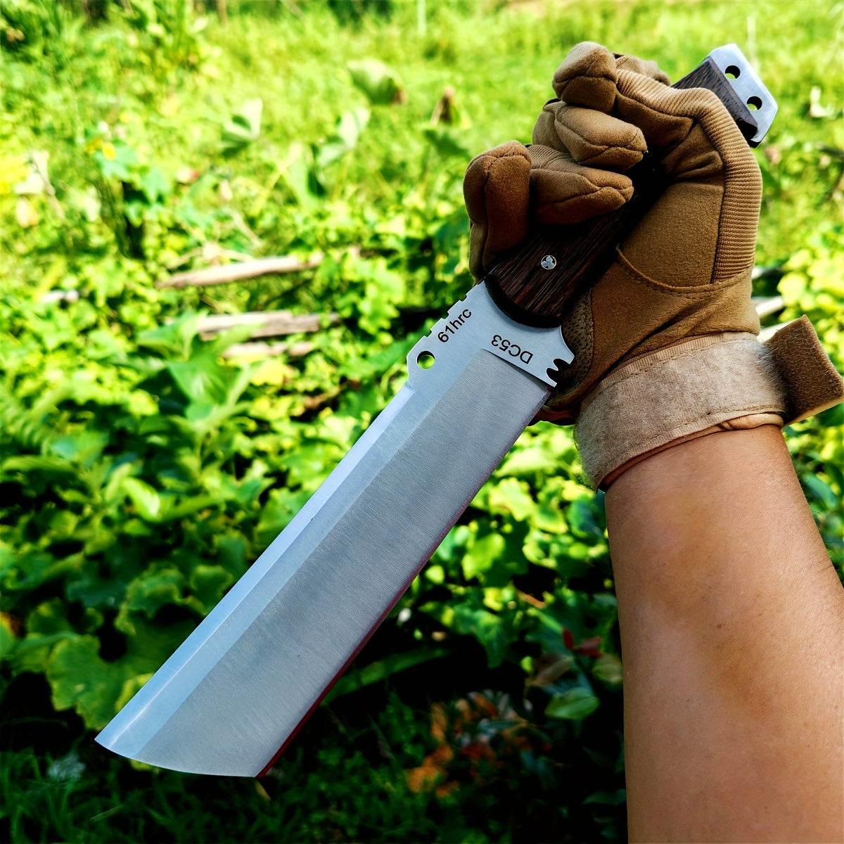Gloved hand holding large survival knife fixed cleaver style blade with wooden handle in green outdoor setting